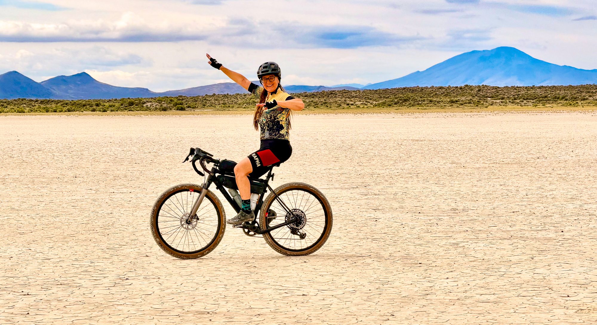 Amy riding hands-free on the playa, arms out wide, mountains behind