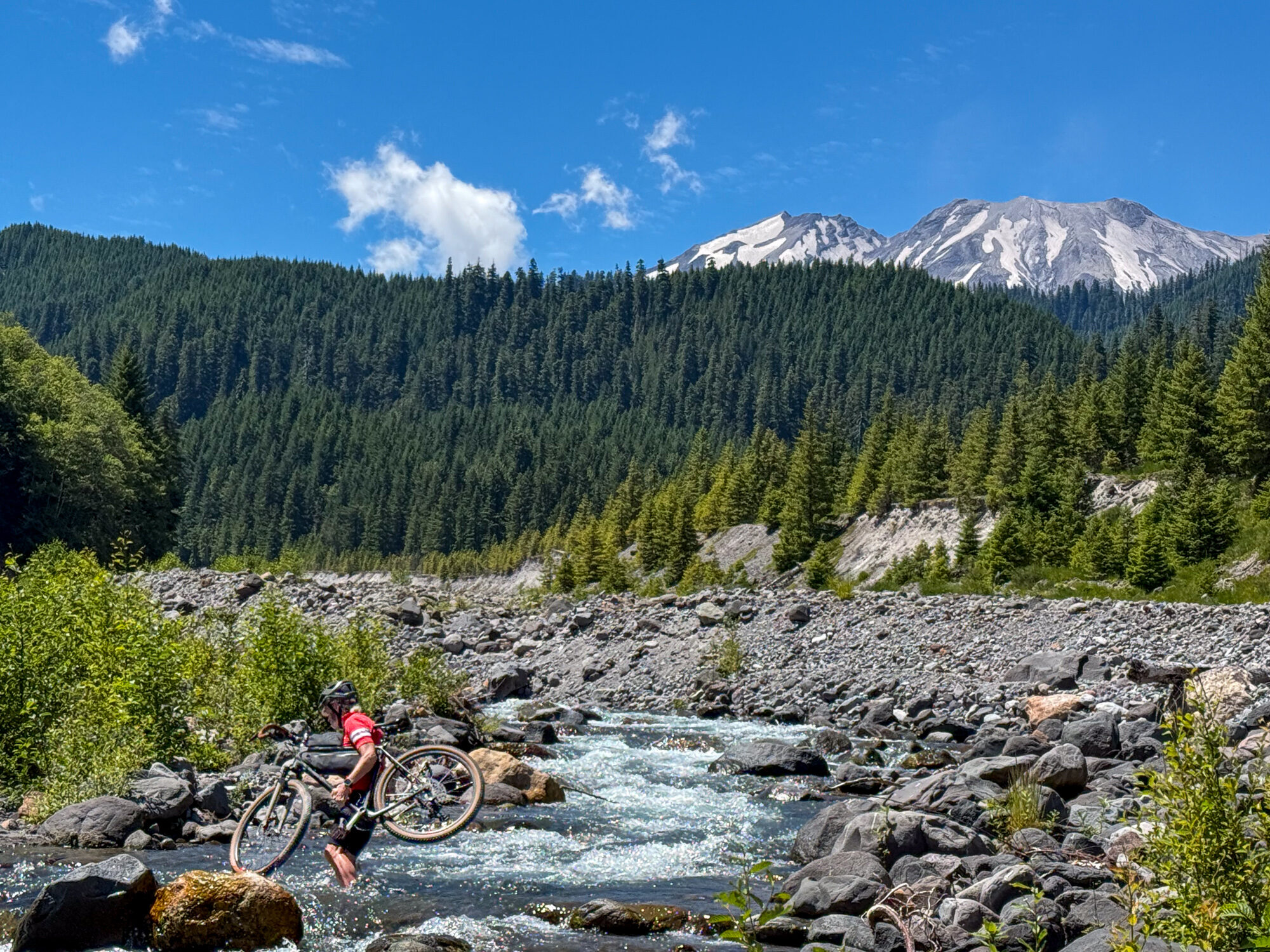 A rider fording beside the cold, fast glacial Muddy River with snowy peaks beyond